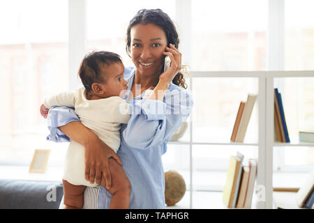 Positive zuversichtlich moderne junge afrikanische Mama stehend gegen Regale und Holding Baby im Arm, während Gespräch am Handy und Kamera Stockfoto