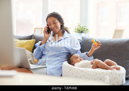 Positive multitasking Junge afrikanische Mutter mit lockigem Haar sitzen auf komfortablen Sofa und schütteln Verlosung für Baby beim Gespräch mit dem Kunden am Telefon Stockfoto