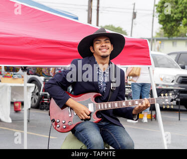 Ein hübscher junger Mann mit dunklen Teint in einem schwarzen, breitkrempiger Hut spielt eine E-Gitarre an der Corpus Christi Southside Farmers' Market. Stockfoto