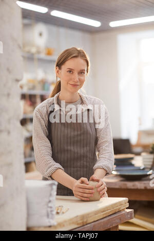 Lächelnd schöne junge Frau in Schürze bei Tisch und Kneten von Ton mit den Händen in der Werkstatt, Sie suchen an Kamera Stockfoto