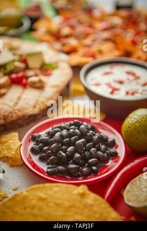 Nahaufnahme auf schwarze Bohnen mit verschiedenen frisch zubereitete Mexikanische Lebensmittel Sortiment. Auf bunten Tisch gelegt. Mit Nachos, Tacos, Tortillas, gegrilltes Fleisch, Dip Stockfoto