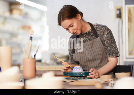 Inhalt attraktive qualifizierte junge Dame in Schürze mit Tabelle und Zeichnung blaue Blätter auf Keramik Schüssel in Töpferei Stockfoto