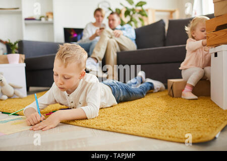 Ernsthafte konzentriert ältester Sohn im casual Outfit liegen auf flauschigen Teppich und Zeichnen mit Bleistift, während seine Schwester finden Spielzeug Stockfoto