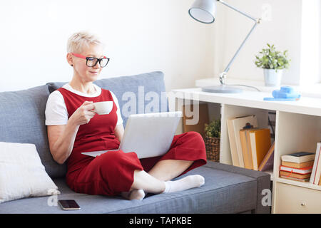 Im mittleren Alter mit überkreuzten Beinen blonde Frau mit Laptop und Tasse Tee auf der Couch sitzen, während Sie Video oder Film Stockfoto