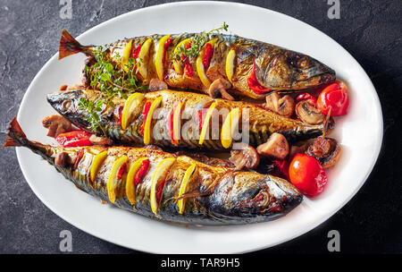 In der Nähe von drei ganze gegrillte Makrelen mit Zitronen, Tomaten, Pilze, Gewürze und Kräuter auf eine weiße, ovale Schale, horizontale Ansicht von oben Stockfoto