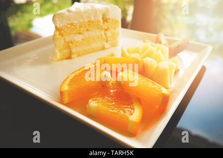 Stück Obst und Milch Kuchen auf weiße Platte/Nahaufnahme von frischen Orange Traube und Apfel Obst Slice auf Tablett mit Kuchen sahne Vanille Dessert auf den Tisch Stockfoto