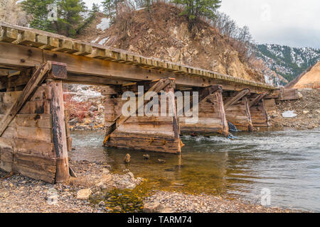 Rustikalen hölzernen Brücke mit Blick auf die grünen Bäume auf einem Berg bei bewölktem Himmel. Die Brücke kreuzen über einen felsigen Stream mit klaren Flachwasser. Stockfoto