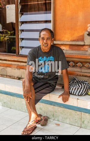 Dusun Ambengan, Bali, Indonesien - 25. Februar 2019: Clan Compound. Nahaufnahme des sitzenden Mann mit orange Wand und Fenster im Rücken. Stockfoto