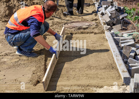 Auf dem Bürgersteig, einem Arbeiter, der reinigt und Ebenen der sandige Plattform mit einem Holzbrett, bereitet die Grundlage für die Festlegung der Pflastersteine rund um Die se Stockfoto