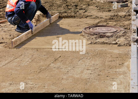 Auf dem Bürgersteig um die Kanalisation Mannloch ein Arbeitnehmer richtet die sandige Plattform mit einem Holzbrett, bereitet die Grundlage für die Festlegung der Pflastersteine. Stockfoto