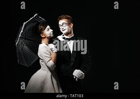 Portrait von Paar mime mit Sonnenschirm auf schwarzen Hintergrund. Mann im Smoking und Gläser und Frau im weißen Kleid. Platz für Text Stockfoto