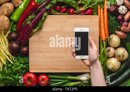 Hand mit Smartphone über Frisches Bio-Gemüse Hintergrund. Veganes Essen Rezepte. Stockfoto