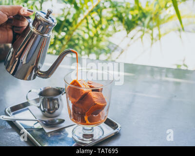 Hand gießen Thai Milch Tee aus Edelstahl Kanne auf Thai Kaffee Eiswürfel im Glas mit Milch in kleinen Kanne auf aus Edelstahl auf Beton Tabelle wi Stockfoto