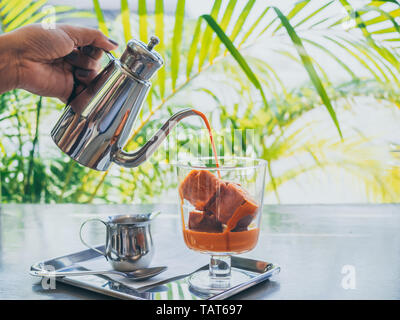 Hand gießen Thai Milch Tee aus Edelstahl Kanne auf Thai Kaffee Eiswürfel im Glas mit Milch in kleinen Kanne auf aus Edelstahl auf Beton. Stockfoto