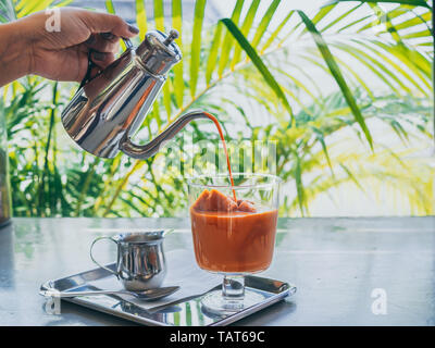 Hand gießen Thai Milch Tee aus Edelstahl Kanne auf Thai Kaffee Eiswürfel im Glas mit Milch in kleinen Kanne auf aus Edelstahl auf Beton. Stockfoto