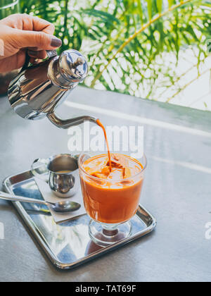 Hand gießen Thai Milch Tee aus Edelstahl Kanne auf Thai Kaffee Eiswürfel im Glas mit Milch in kleinen Kanne auf aus Edelstahl auf Beton Tabelle ve Stockfoto