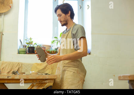 Konzentriert bärtigen Kerl. Dunkelhaarige erfahrenen Mann in schutzschürze Holding vorbereitetes Material während der Arbeit Stockfoto