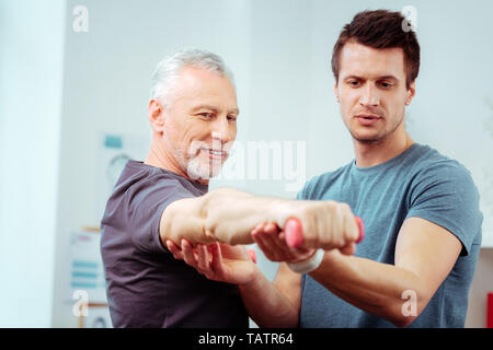 Ich fühle mich stark. Freudige alter Mann auf seine Hand mit einer Hantel während Gefühl Stärke in Es Stockfoto