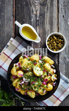 Köstliche neue Kartoffel Salat mit roten Zwiebeln, Kapern, Grüns in einem schwarzen Schüssel. Senf Essig Dressing und Kapern auf einem alten Holz- rustikalen Tisch, vertikal Stockfoto