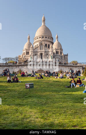 Paris, Frankreich, 12. April 2019: Leute sitzen auf der Wiese vor der Basilika Sacré-coeur auf dem Hügel Montmartre an einem sonnigen Tag Stockfoto