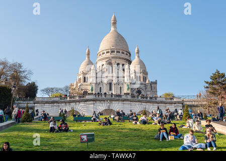 Paris, Frankreich, 12. April 2019: Leute sitzen auf der Wiese vor der Basilika Sacré-coeur auf dem Hügel Montmartre an einem sonnigen Tag Stockfoto