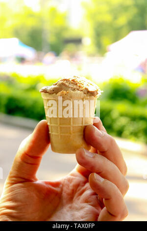 Eine männliche Hand Schokolade Eis in der Waffel Kegel mit unscharfen grüne Natur Hintergrund. Sonnenschein bei Tag und Blick in die Natur. Selectuve soft Fokus. Tex Stockfoto