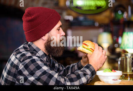 Pub ist ein entspannender Ort Drink zu entspannen. Mann mit Bart Bier trinken essen burger Menü. Essen im Pub. Brutale hipster bärtigen Mann an der Theke sitzen. Kalorienreiche Snacks. Hipster entspannen im Pub. Stockfoto