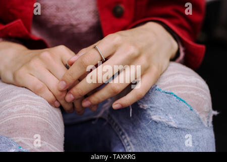 Frau sitzt gekreuzten Beinen mit den Händen in der Runde warten. Zerrissene Jeans und rote Jacke. Berühren Hände besorgt/Sad Stockfoto