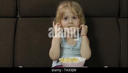Schöne kleine blonde Mädchen auf einem Sofa zu Hause sitzen und essen Mais Puffs. Niedliche kind Geschmack puffcorns. Im Inneren. Kinder essen Stockfoto
