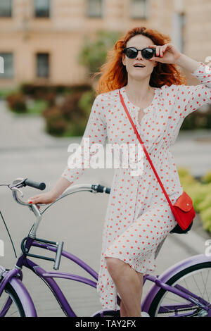 Street Style und aktive Erholung. Curly rothaarige Frau hält die Hand auf Frame Sonnenbrille, trägt weiße Kleid, in die Ferne gerichtet, wartet auf fr Stockfoto