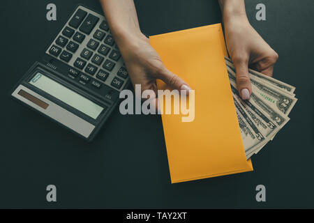 Das Mädchen hält ein gelber Umschlag mit Geld auf dunklem Leder Hintergrund, auf dem Tisch ist ein Taschenrechner. Konzept der Gehalt oder Bestechung. Ansicht von oben. Stockfoto