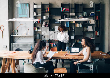 Drei junge multikulturelle Kollegen diskutieren Projekt in modernen Loft - Einfallsreich, Interaktion, Engagement Stockfoto