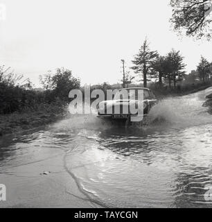 1960, historische, ein Aston Martin Sportwagen erstellt einen splash Wie geht es durch das Wasser auf einem überfluteten Landstraße, Buckinghamshire, England, UK. Stockfoto