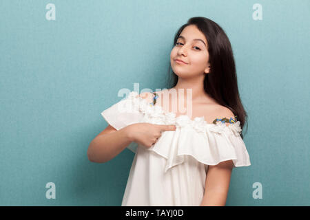 Dies ist mir. Portrait von Stolz schöne Brünette junge Mädchen mit schwarzen glatten Haaren in weißem Kleid stehend, sich zeigen und auf Kamera. Stockfoto