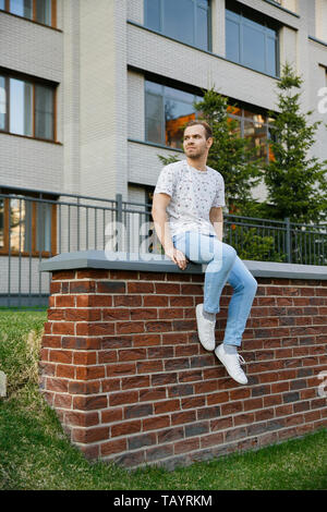 Junge schöne stilvolle Mann mit Borsten sitzen auf einem gemauerten Zaun in der Nähe der Gebäude an einem Sommerabend bei klarem Wetter Stockfoto