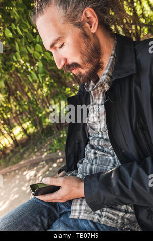 Junge Bartgeier lächelnden Mann mit einem Smartphone sitzt in einem Sommer Park, vertikal Outdoor Portrait Stockfoto