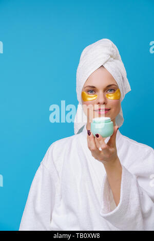 Das Bild der jungen Frau mit Gel Pads unter den Augen und ein Handtuch auf dem Kopf und mit Sahne in der Hand auf leeren blauen Hintergrund Stockfoto