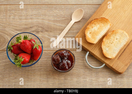 Traditionelle hausgemachte Erdbeermarmelade in eine Glasschüssel, frische Beeren, Toast auf Schneidebrett mit Löffel auf hölzernen Schreibtisch. Ansicht von oben. Stockfoto