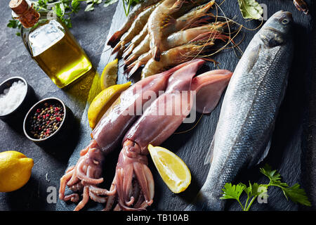 Wolfsbarsch, Garnelen, Tintenfisch, Zitronenscheiben, Gewürze und Kräuter auf einer Schiefertafel Platte auf einer konkreten Tabelle, Ansicht von oben, close-up Stockfoto