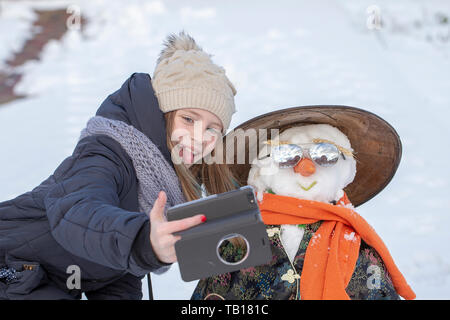 Adorable junges Mädchen ist, die Bilder von selfie mit einem Schneemann in schönen Winter Park. Winter Aktivitäten für Kinder. Nahaufnahme Stockfoto