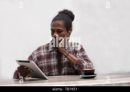 Eine junge schwarze Mann konzentriert mit Tablet und Kaffee, ein papua junger Mann mit Tablette mit einer Tasse Kaffee Stockfoto