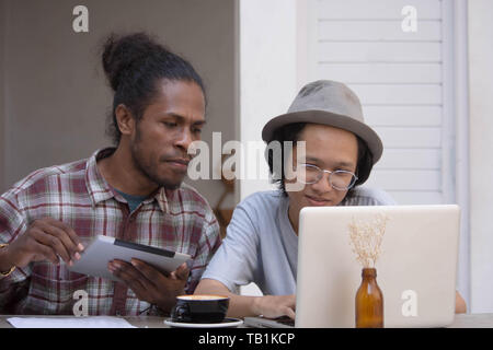Zwei junge kreative Menschen mit Laptop und Tablet diskutieren, jungen asiatischen und schwarzer Mann arbeiten mit Tablet-PC und Laptop in ein Cafe, Papua und asiatischer Mann arbeiten Stockfoto