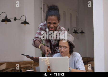 Zwei junge kreative Menschen mit Laptop und Tablet diskutieren, jungen asiatischen und schwarzer Mann arbeiten mit Tablet-PC und Laptop in ein Cafe, Papua und asiatischer Mann arbeiten Stockfoto
