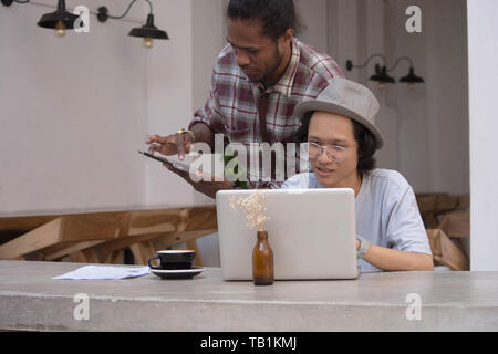 Zwei junge kreative Menschen mit Laptop und Tablet diskutieren, jungen asiatischen und schwarzer Mann arbeiten mit Tablet-PC und Laptop in ein Cafe, Papua und asiatischer Mann arbeiten Stockfoto