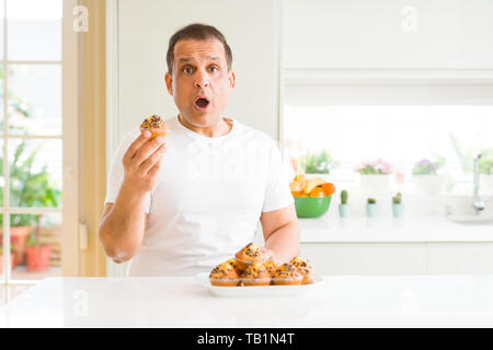 Mittleres Alter Mann essen Chocolate Chip Muffins zu Hause Angst im Schlag mit einer Überraschung konfrontiert, ängstlich und aufgeregt mit Angst Ausdruck Stockfoto
