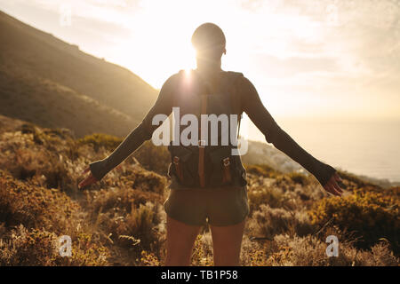 Ansicht der Rückseite Frau mit Rucksack auf Land weg an der Sonne mit ihren Hände ausgestreckt. Weibliche Wanderer Umarmung der Sonne Licht. Stockfoto