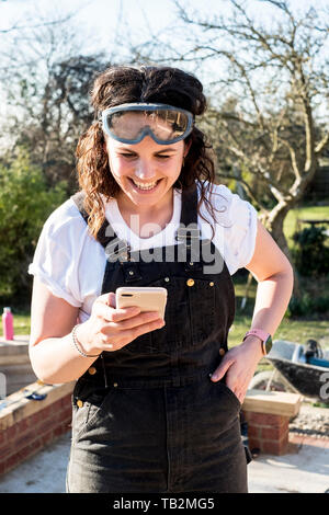 Lächelnde Frau mit Latzhose und Schutzbrille ihr Handy prüfen. Stockfoto
