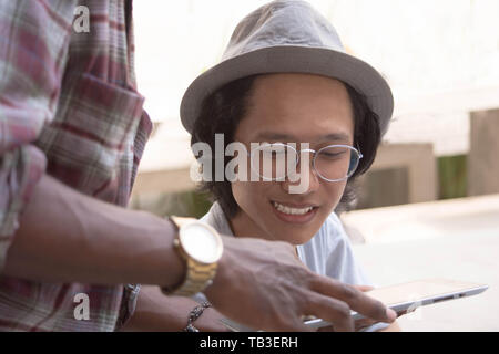 Jungen asiatischen Mann mit Hut und Brille lächelnd während sie etwas mit Freund mit einer Tablette diskutieren Stockfoto