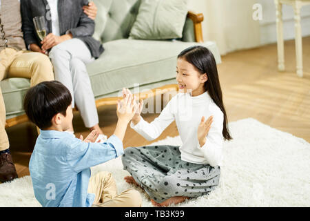Zwei süße kleine asiatische Kinder Bruder und Schwester saß auf dem Teppich spielen Spiel mit Eltern sitzen auf einem Sofa im Hintergrund Stockfoto