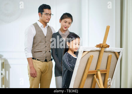 Schöne kleine asiatische Mädchen mit langen schwarzen Haaren und einem Gemälde auf Leinwand, während die Eltern hinter dem Ständigen beobachten. Stockfoto